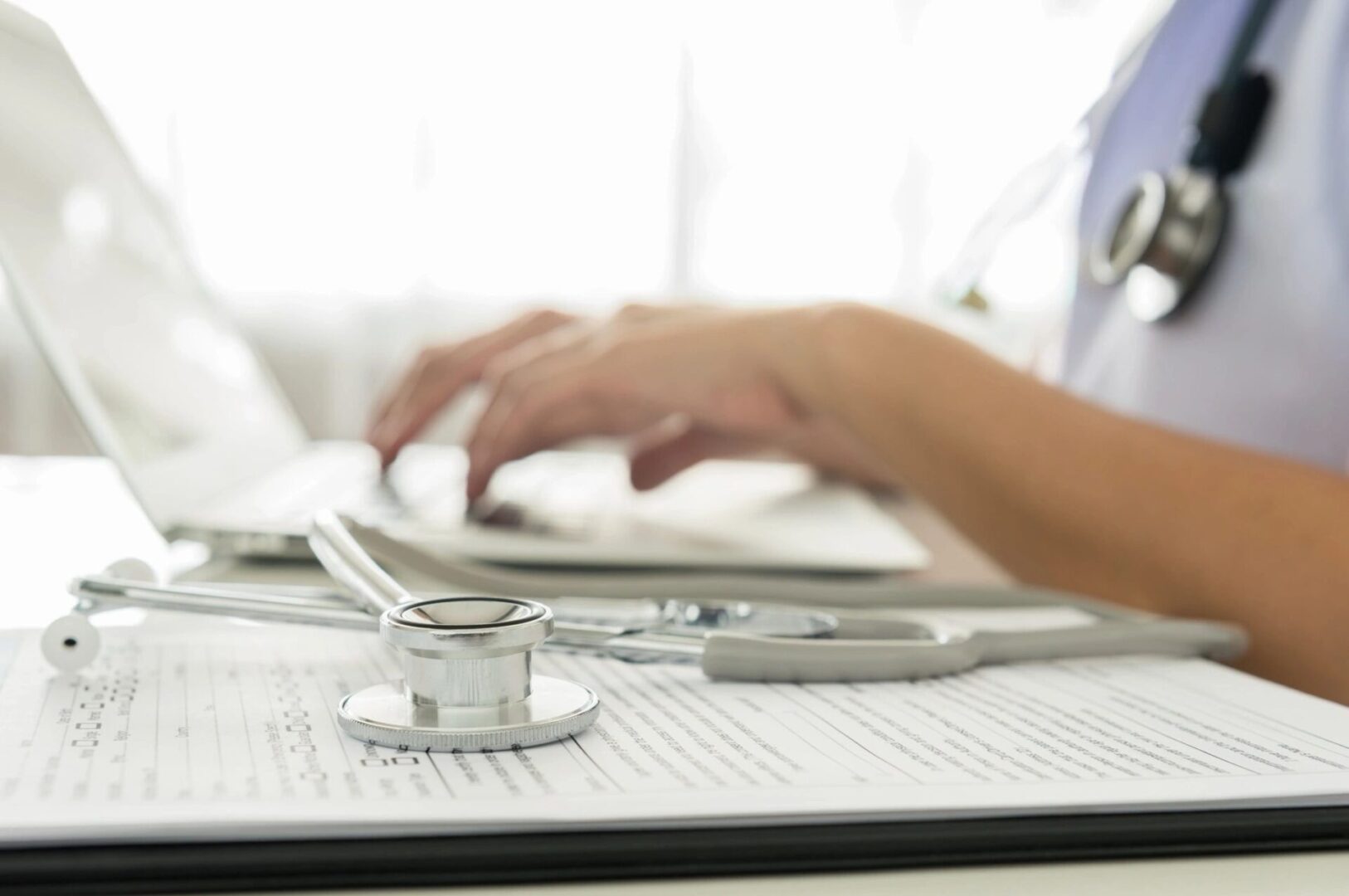 A person is typing on a laptop with a stethoscope around it.