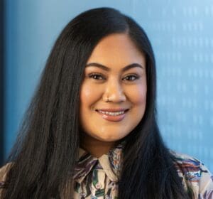 A woman with long black hair smiling, wearing a multicolored shirt, standing indoors with a blue patterned background.