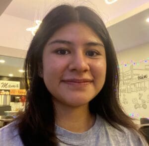 Close-up of a smiling young woman with long dark hair in a blue t-shirt, indoors with soft lighting and a blurred restaurant background.