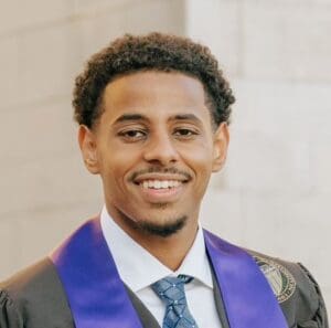 A young man in a graduation gown and tie smiles confidently at the camera.