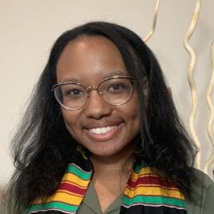 A smiling woman with glasses, wearing a green blouse and a colorful striped scarf, stands in front of a neutral background.