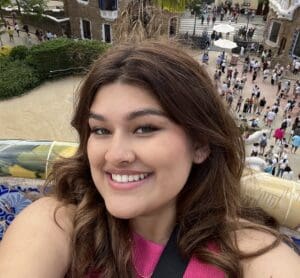 Smiling woman with curly hair and a pink top, taking a selfie, with a busy park scene in the background.