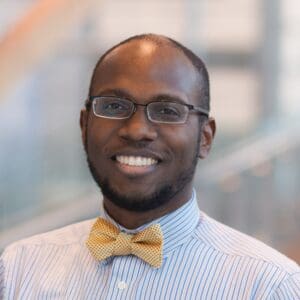 A portrait of a smiling black man wearing glasses and a bow tie, standing indoors with a blurred background.