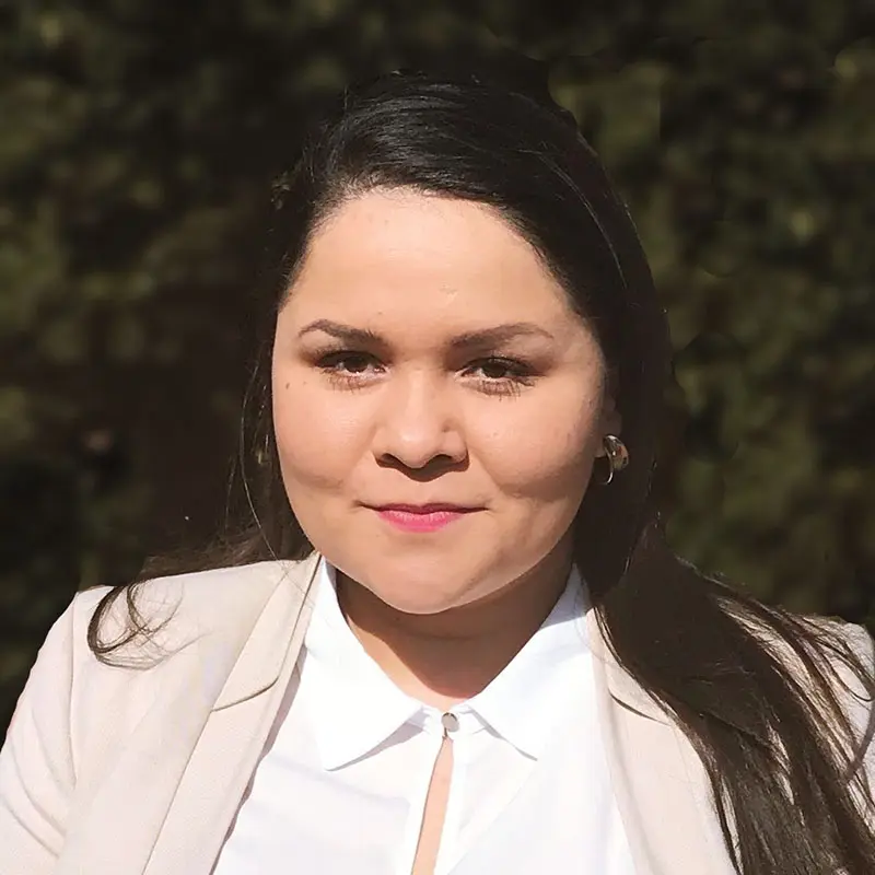 A woman with long dark hair and a light-colored blazer stands outside, facing the camera with a neutral expression.