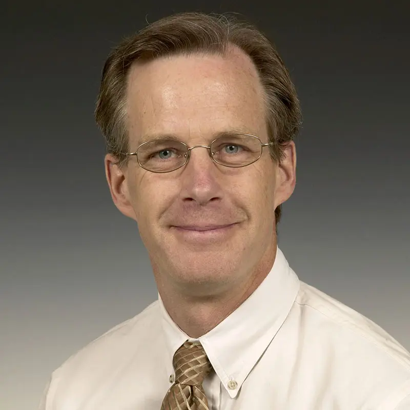 A man with short hair, wearing glasses, a white shirt, and a beige-striped tie, smiles at the camera against a gray background.