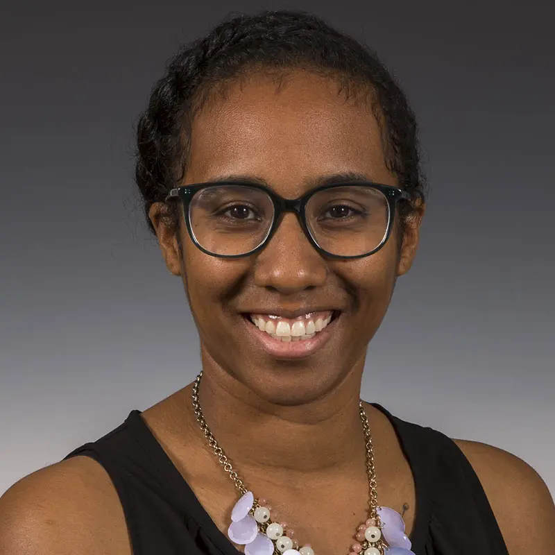 A person with curly hair, wearing glasses, a black sleeveless top, and a necklace, smiles at the camera with a gray gradient background.