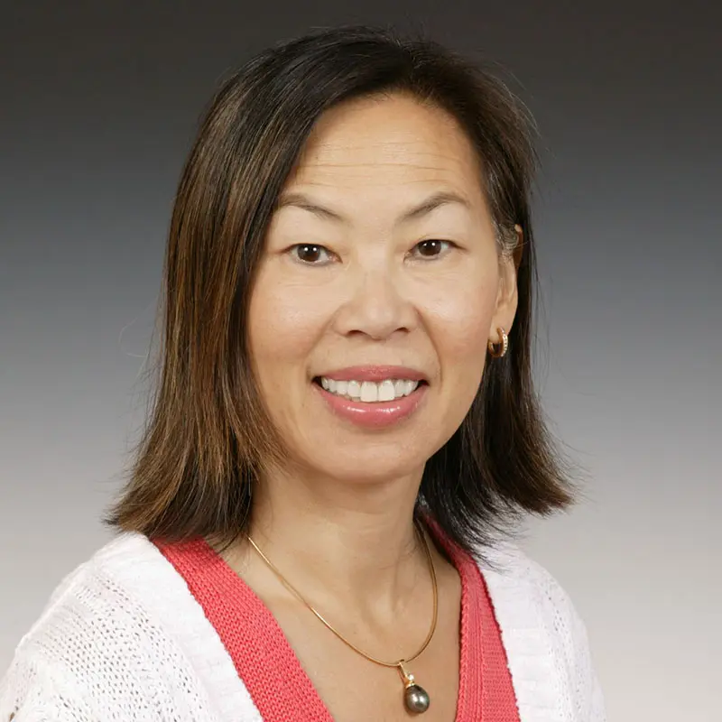 A woman with shoulder-length hair smiles, wearing a white and pink top and a gold necklace with a pearl pendant. She has gold earrings and stands against a plain background.