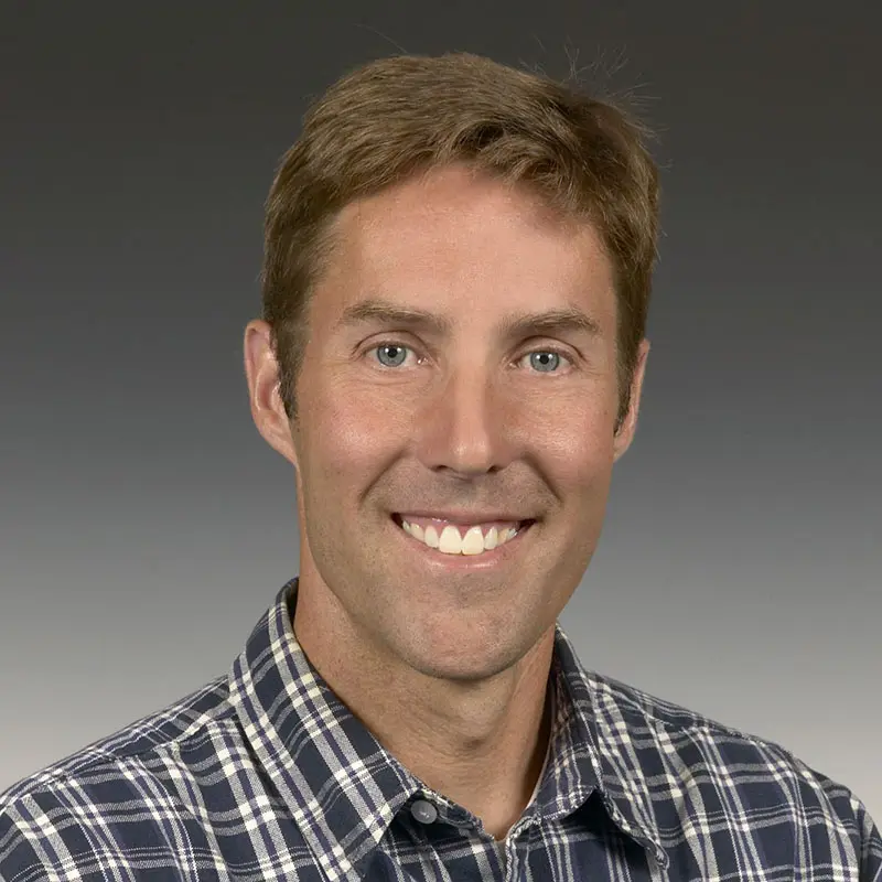 A man with short brown hair is smiling. He is wearing a blue and white checkered shirt. The background is plain and gradiented from dark to light gray.