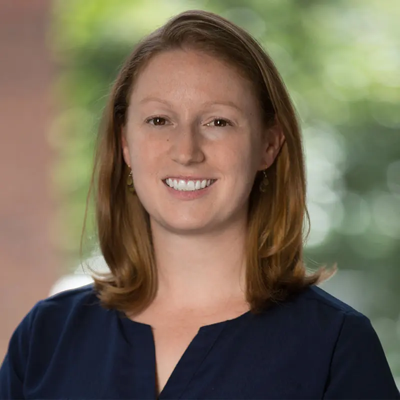 A woman with shoulder-length red hair smiles at the camera. She is wearing a navy blue top, and the background is blurred greenery.