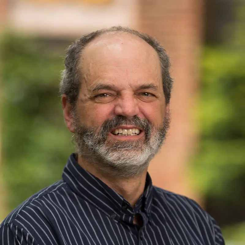 A man with a bald head and grey beard is smiling, wearing a dark striped shirt. A blurred green and brick background is visible.