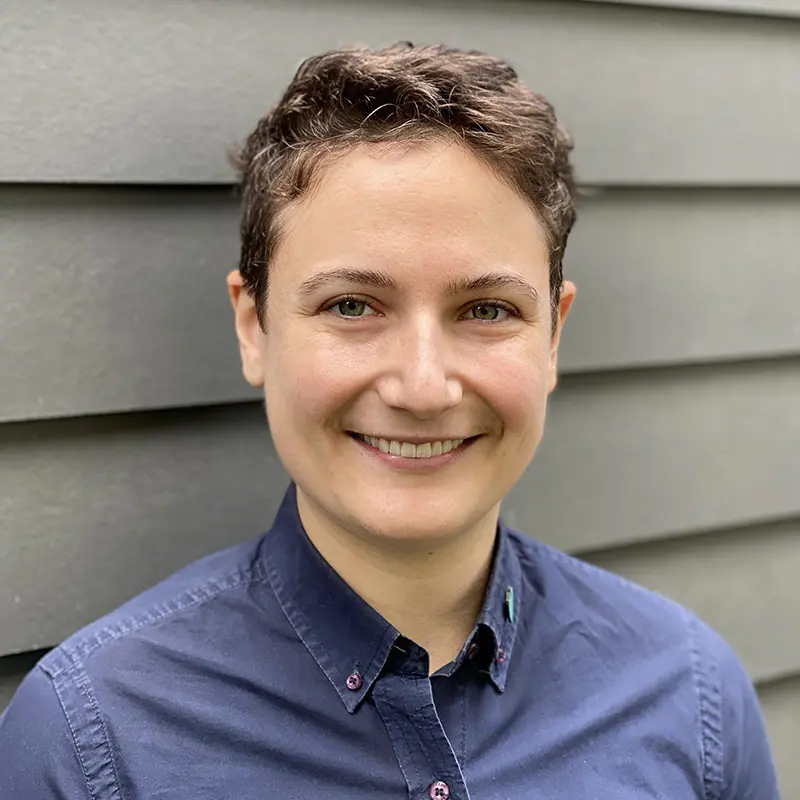 Person with short hair and a blue button-up shirt smiling, standing in front of a gray, horizontally paneled wall.