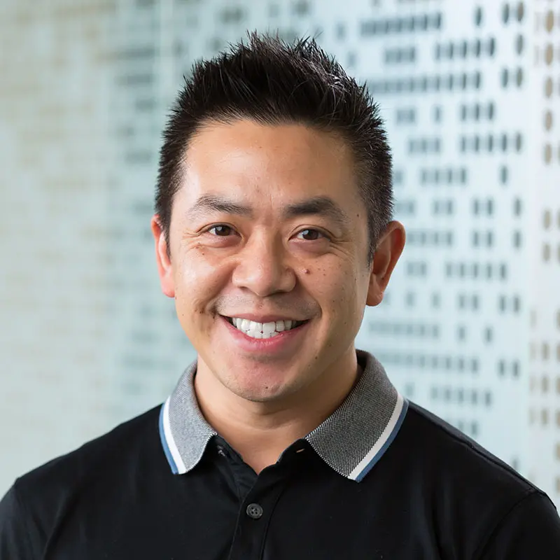 A man with short, spiky hair and wearing a dark polo shirt with a light grey collar smiles at the camera. The background features blurred text or symbols on a light surface.