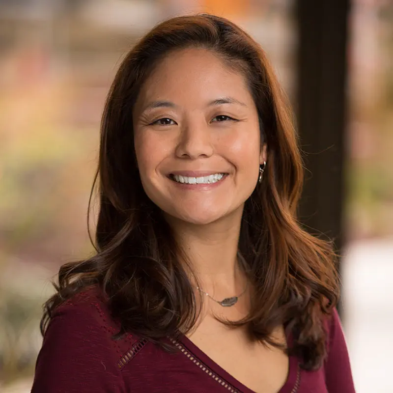 A woman with long brown hair wearing a maroon top is smiling at the camera with a blurred background.