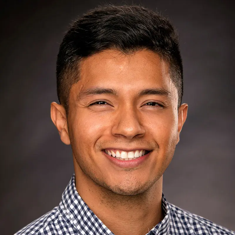 A man with short dark hair smiles at the camera against a dark background, wearing a checkered shirt.