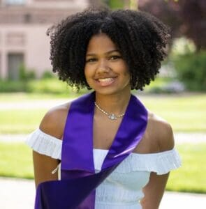 A young woman with curly hair wearing a white off-shoulder dress and a purple graduation stole stands outdoors, smiling at the camera.