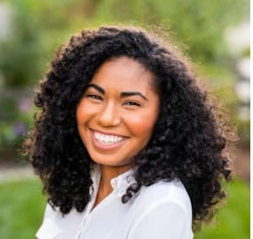 A woman with curly black hair smiles outdoors, wearing a white shirt. The background is blurred greenery.