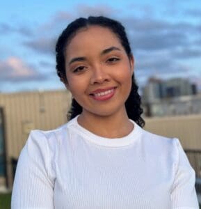 A woman with curly hair wearing a white top smiles outdoors with a building and sky in the background.