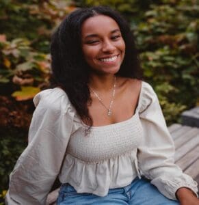 A woman with long dark hair wearing a white blouse and jeans sits outdoors on a wooden bench, smiling at the camera with greenery in the background.
