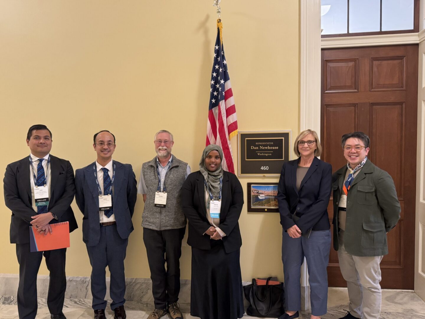 Six people stand in a row in front of an office door, a wall plaque, and an American flag, posing for a group photo in a hallway.