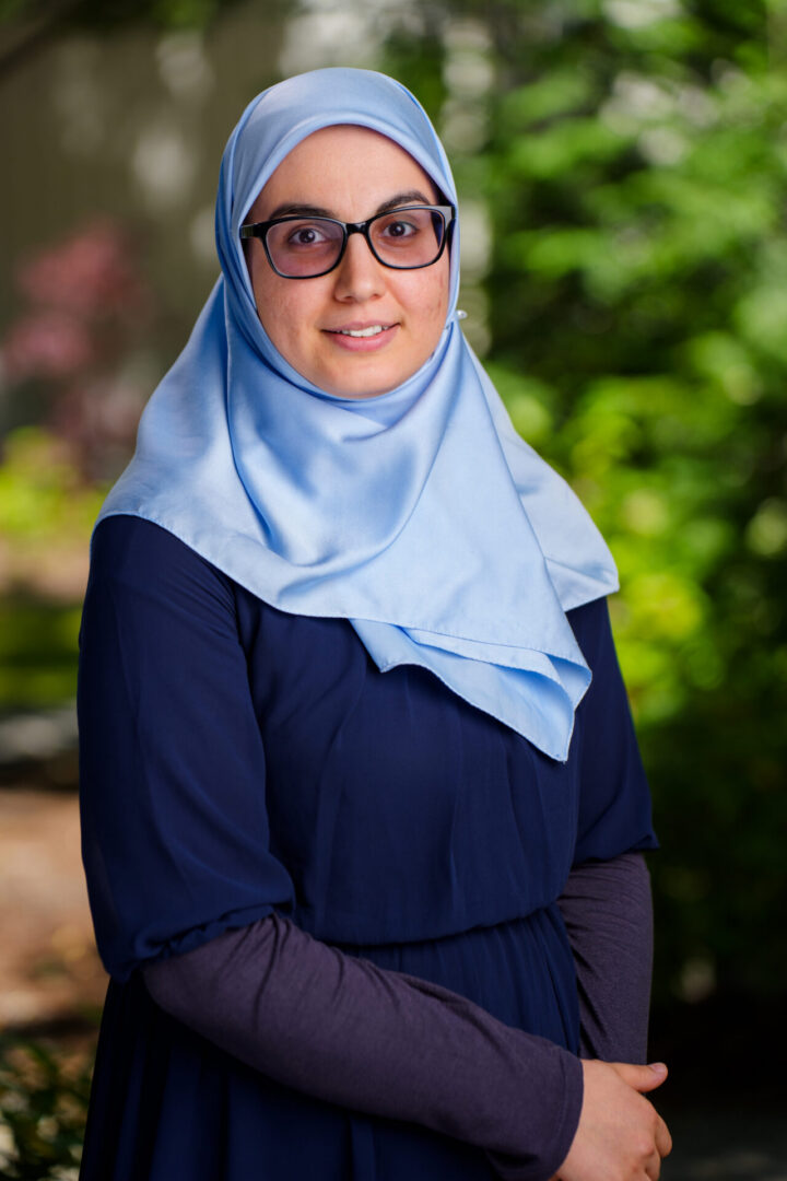 A woman wearing glasses, a light blue hijab, and a navy blue dress stands outdoors with greenery in the background.