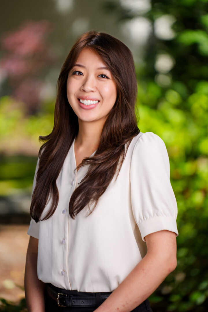 A woman with long brown hair wearing a white blouse stands outdoors, smiling at the camera with greenery in the background.